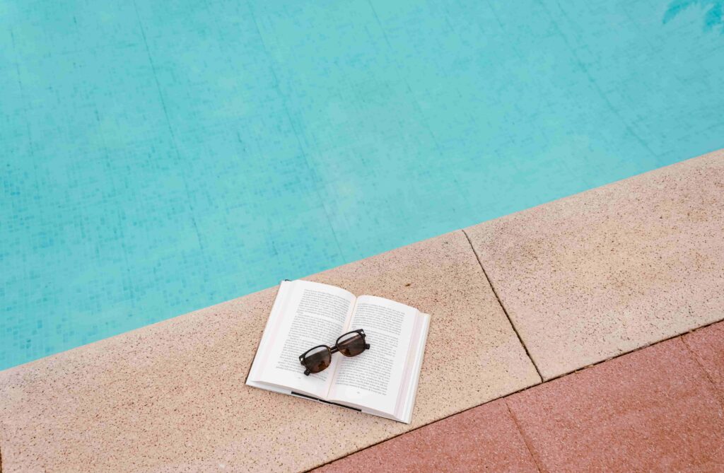 libro abierto con gafas de sol en el borde de una piscina azul